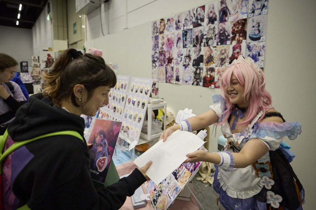 Foto aus der Artist Alley der POLARIS Convention 2024. Man sieht wie eine Dame im Cosplay eine Zeichnung an eine Kundin überreicht.