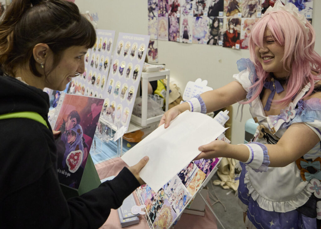 Foto aus der Artist Alley auf der POLARIS Convention 2024. Eine Künstlerin gibt einer Kundin ein Kunstwerk