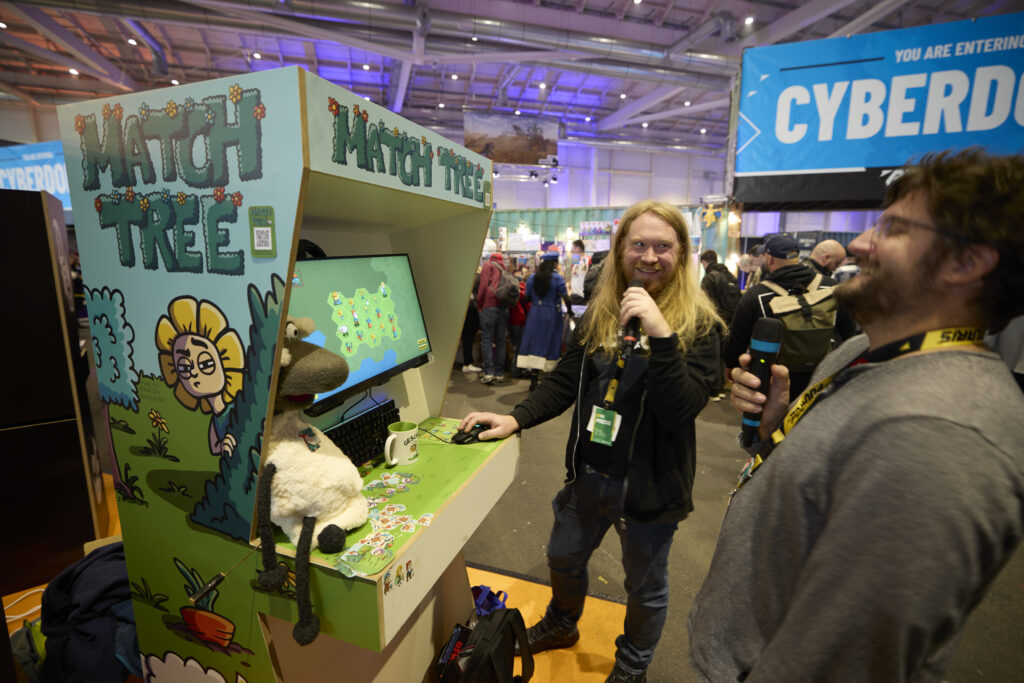 Foto aus der Indie Area der POLARIS Convention. Zwei Aussteller:innen stehen vor einer Arcade Booth des Spiels MATCH TREE.