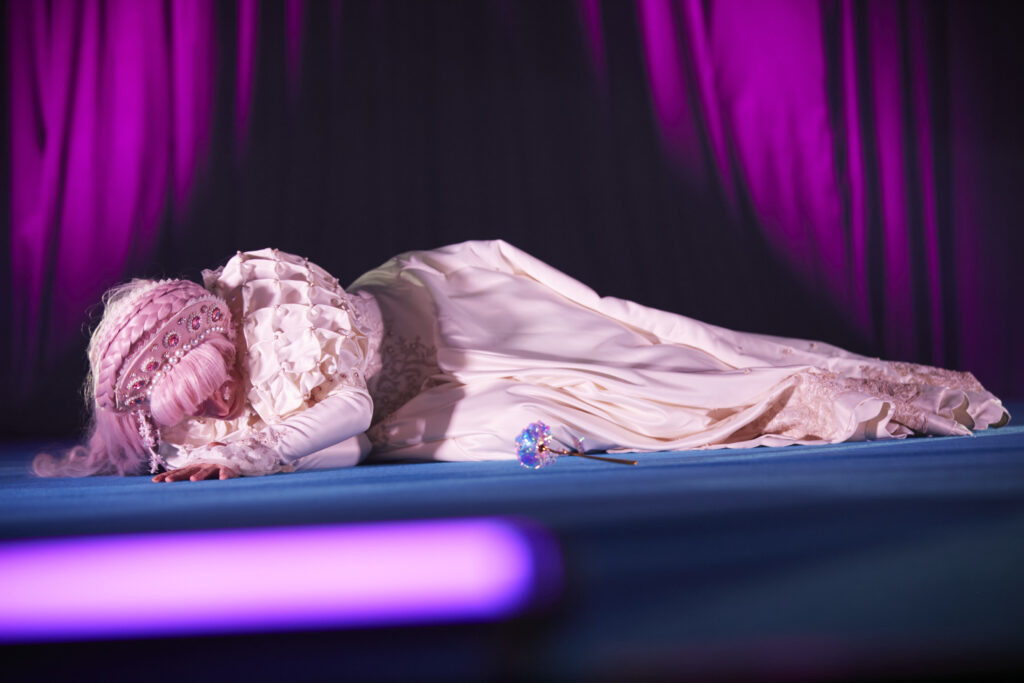 Foto einer Cosplayerin, die während ihrer Performance im Cosplaywettbewerb auf dem Boden liegt. Vor ihr liegt eine künstliche Blume und sie trägt ein weißes, gerüschtes Kleid sowie ein detailliertes Haarband.